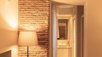 Un pasillo con pared de ladrillo y una lámpara. Una puerta da a un baño con lavamanos y espejo.