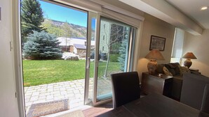 Una habitación con puerta de vidrio corrediza que da a un patio trasero con césped, un árbol y una parrilla para barbacoa. Hay una mesa con sillas y una lámpara en una mesita auxiliar.