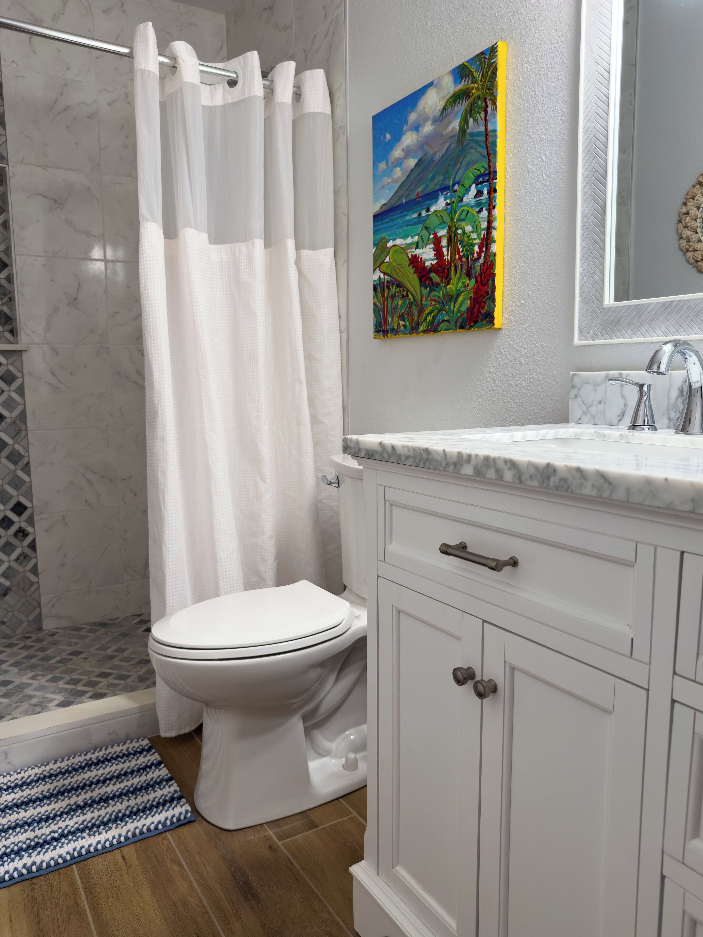 Une salle de bain avec des toilettes blanches, un rideau de douche, un lavabo avec un miroir et un tableau représentant un paysage tropical.