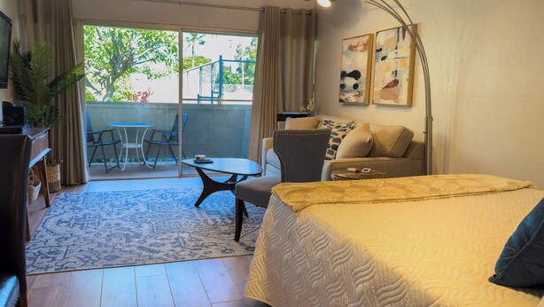 A hotel room with a bed, a desk, a chair, a table, and a balcony with a view of greenery. - Newly remodeled studio, jacuzzi and pool (Kihei)