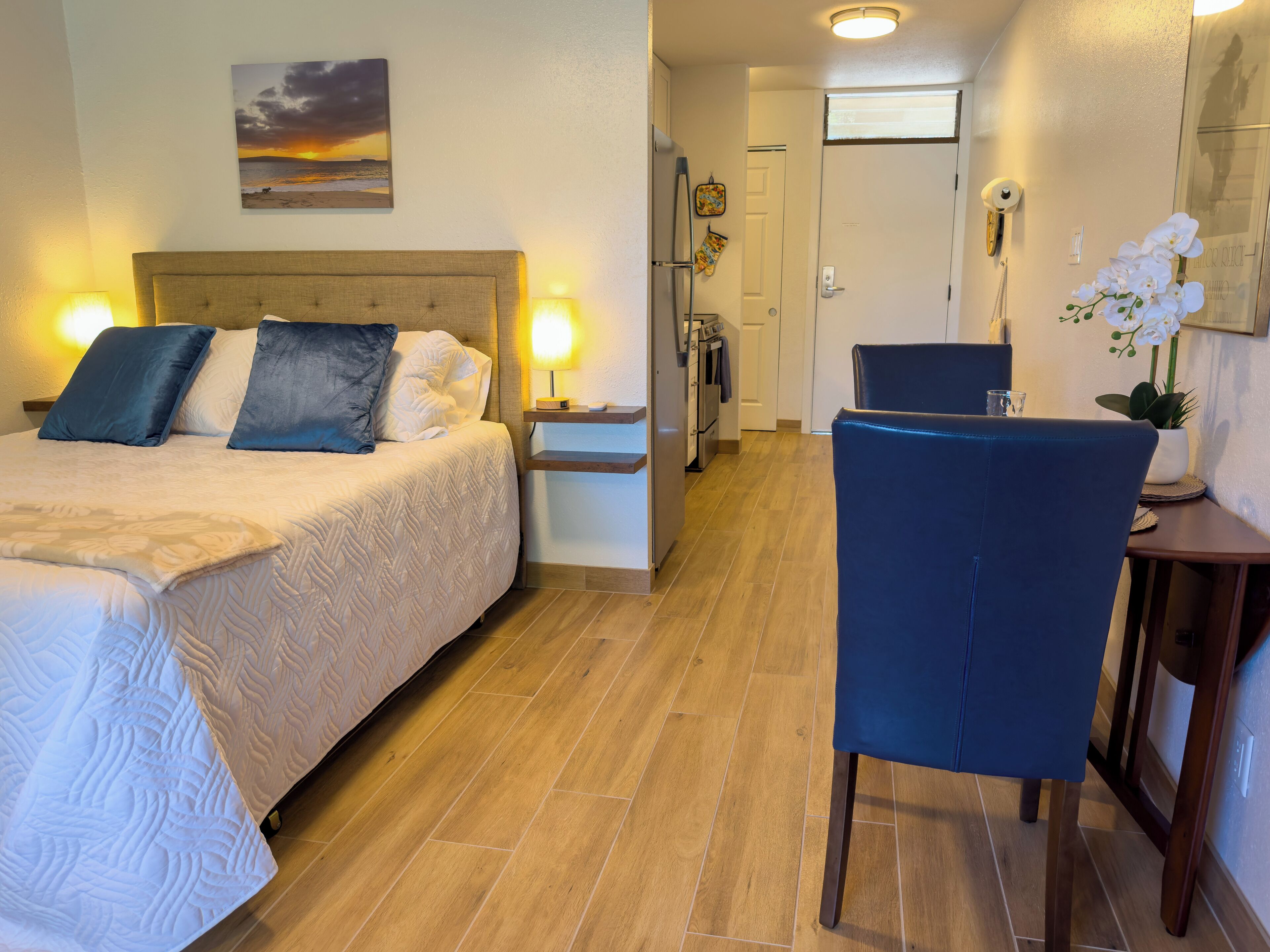 Una camera d'albergo con un letto, lampade sui comodini, una scrivania con una sedia, un tavolino con un vaso di fiori e un angolo cottura.