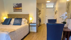 A hotel room with a bed, bedside lamps, a desk with a chair, a small table with a vase of flowers, and a kitchen area. - Newly remodeled studio, jacuzzi and pool (Kihei)