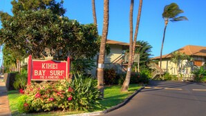A sign for 'KIHEI BAY SURF' with a clear view of the street, palm trees, and a building with a red roof.