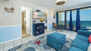 A living room with a sofa, a dining table with chairs, a TV, and a view of the sea through large windows. - BEACH FRONT! Condo in HIGH Demand! (Gulf Shores)