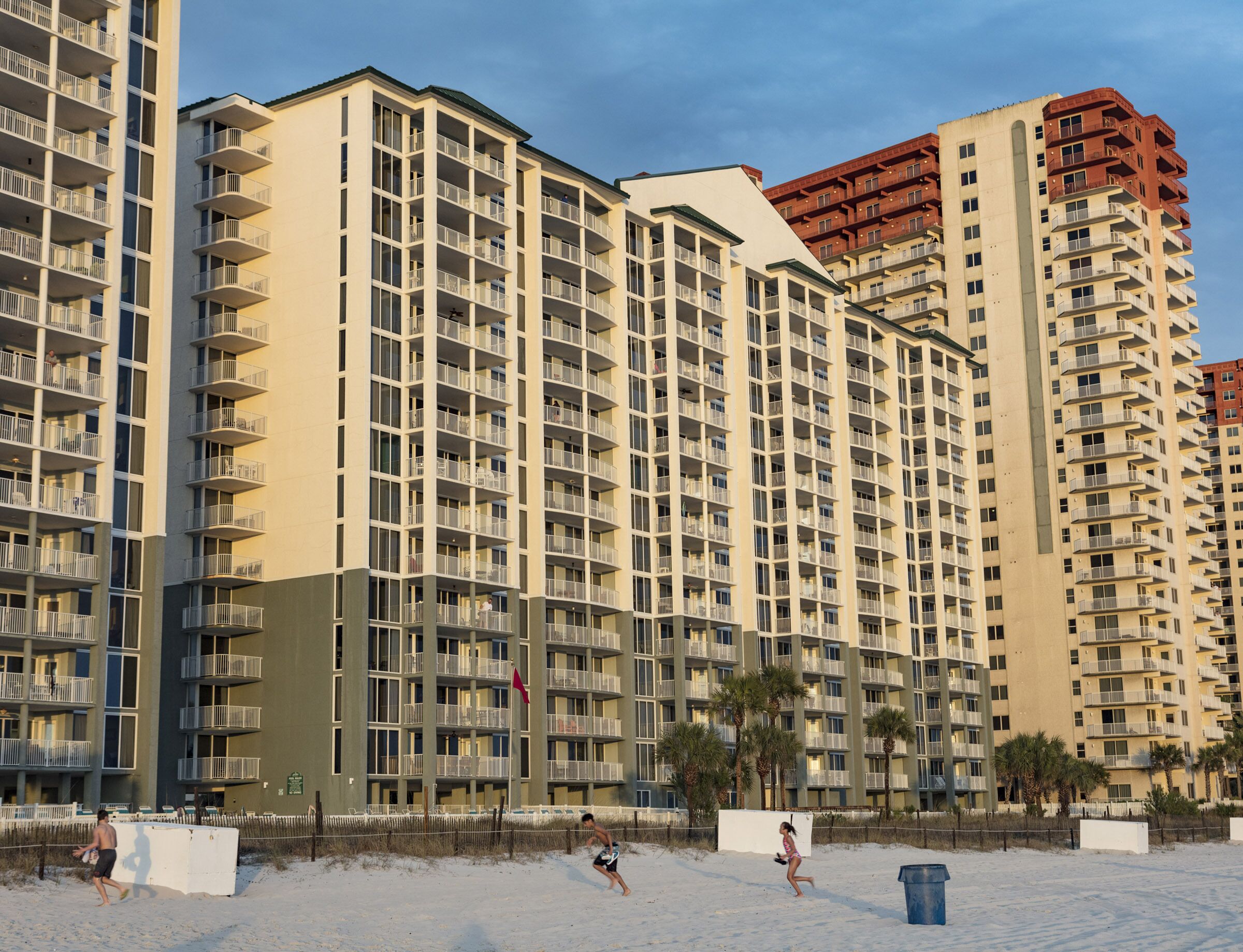A high-rise hotel with multiple balconies, a sandy beach in front, and palm trees.