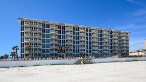 Un hôtel à plusieurs étages doté de balcons, situé en bord de plage, avec une plage de sable fin devant et des palmiers.