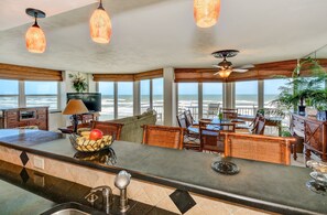 A spacious living area with a view of the ocean, featuring a kitchen area with a counter and sink, wicker furniture, and pendant lighting.