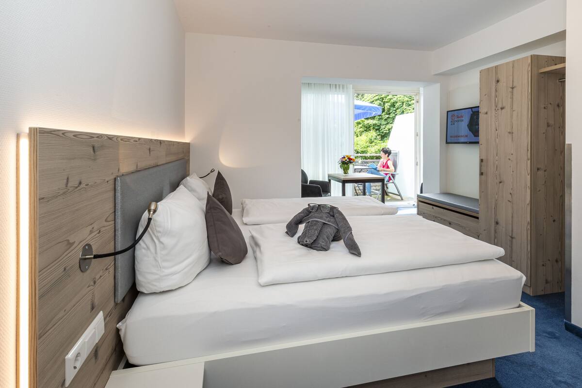 A modern hotel room with a wooden headboard, a bed with white linens, a bedside table, and a window with a view of the outdoors.