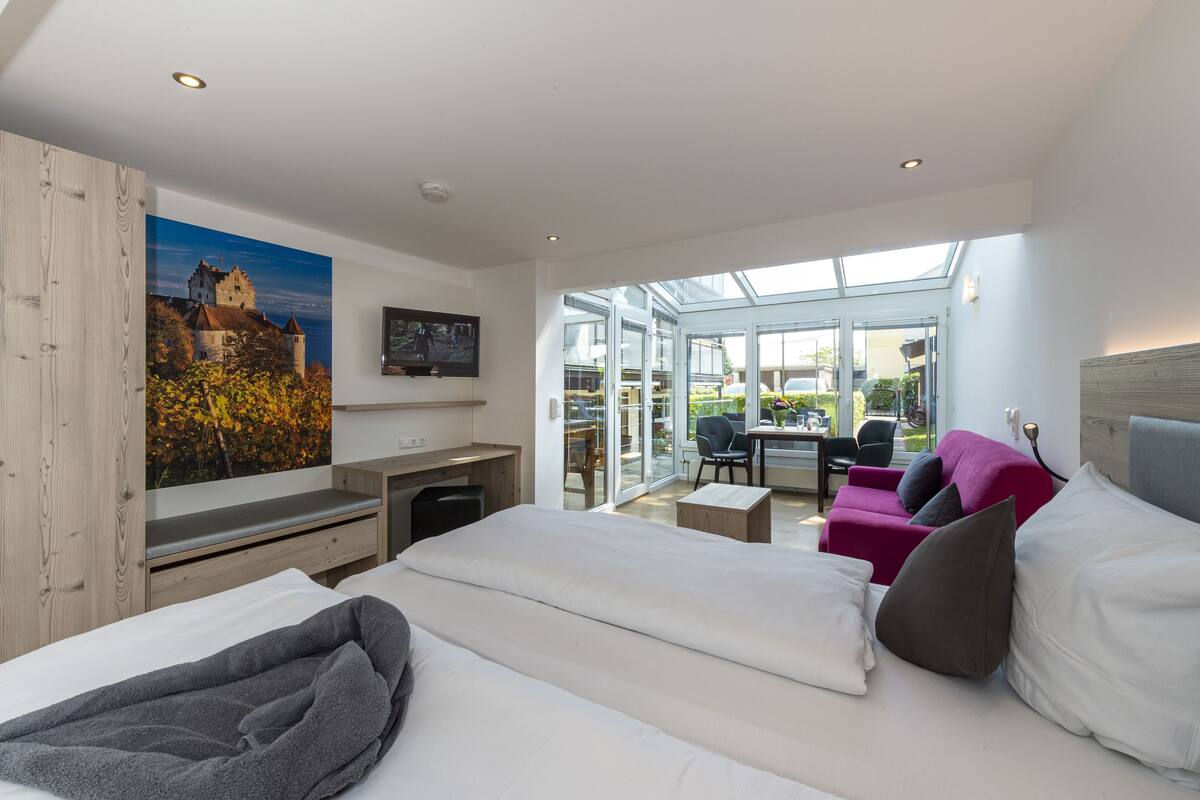 A modern hotel room with a large bed, a desk, a chair, and a sofa. There is a large window providing a view of the outside.