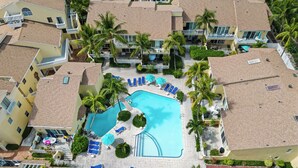 Eine Vogelperspektive zeigt ein Resort mit einem zentral gelegenen Schwimmbad, umgeben von Liegestühlen und Palmen.