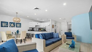 Ein modernes Wohnzimmer mit einer Couch, Sesseln und einem Couchtisch. Im Hintergrund ist eine Küchenzeile mit weißen Schränken und einem Esstisch zu sehen.