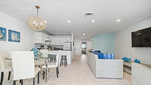 Eine moderne Küche mit weißen Schränken, ein Essbereich mit einem Glastisch und ein Wohnzimmer mit einem weißen Sofa und blauen Kissen.