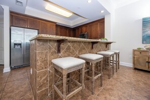 Una cocina con encimera de granito, refrigerador de acero inoxidable, armarios de madera y una zona con taburetes de bar.