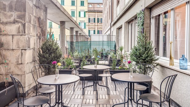 Una terraza con mesas y sillas redondas, rodeada de plantas en macetas y vistas a edificios.