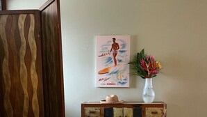 Una stanza con un letto, un armadietto in legno con cassetti, un poster di un surfista e un vaso con fiori.