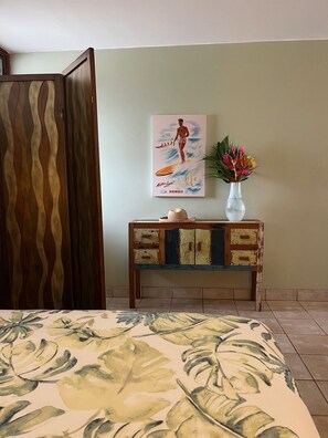 Una habitación con cama, un mueble de madera con cajones, un póster de un surfista y un florero con flores.