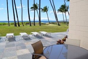 An outdoor patio with a glass table and chairs, overlooking a grassy area with palm trees and the ocean. - Ocean front ground floor condo. HKOK 109 (Kihei)