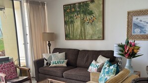 Un soggiorno con divano, tavolino in vetro, ventilatore a soffitto, un quadro e una finestra con tende.