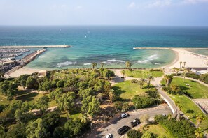 An aerial view of a coastal area with a marina, beach, and park with trees and pathways.