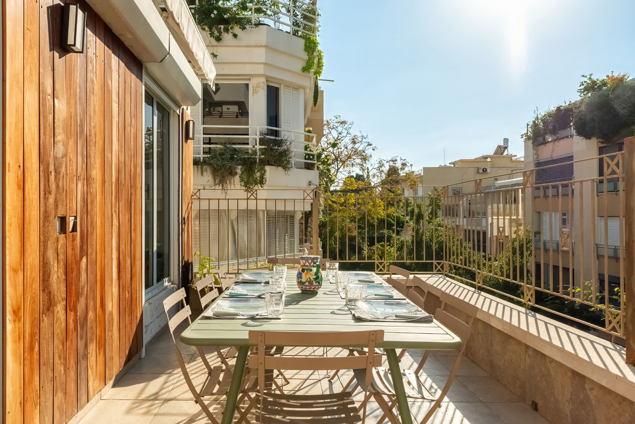 Un balcón con una mesa lista para dos, una puerta de madera, y un edificio con balcones y follaje.
