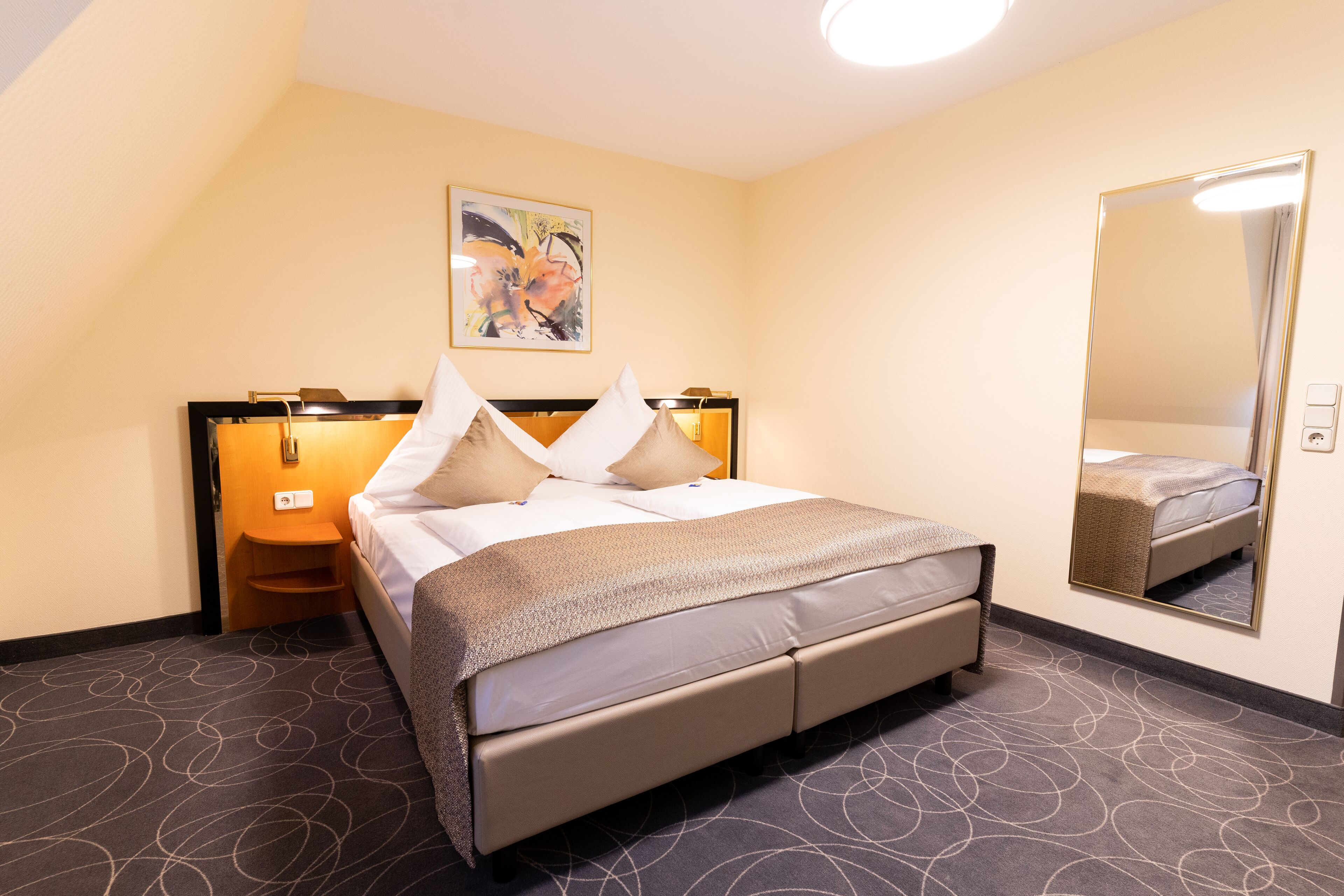 A hotel room with a bed, a nightstand, a wall-mounted mirror, and a painting on the wall.