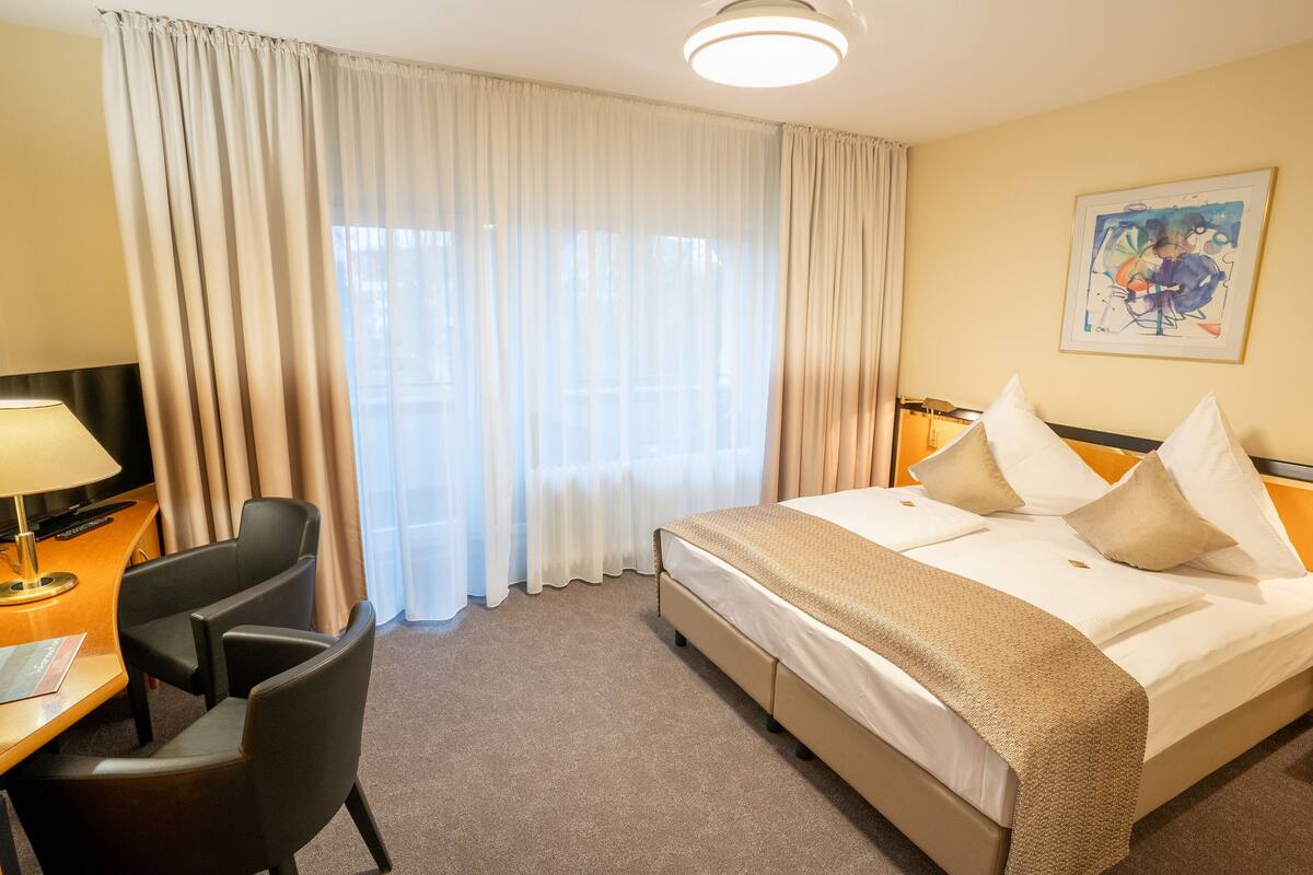 Quarto de hotel com cama, escrivaninha, luminária, cadeira e uma grande janela com cortinas.