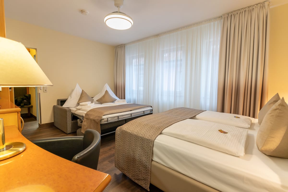 Quarto de hotel com cama, escrivaninha, cadeira e uma janela grande com cortinas.