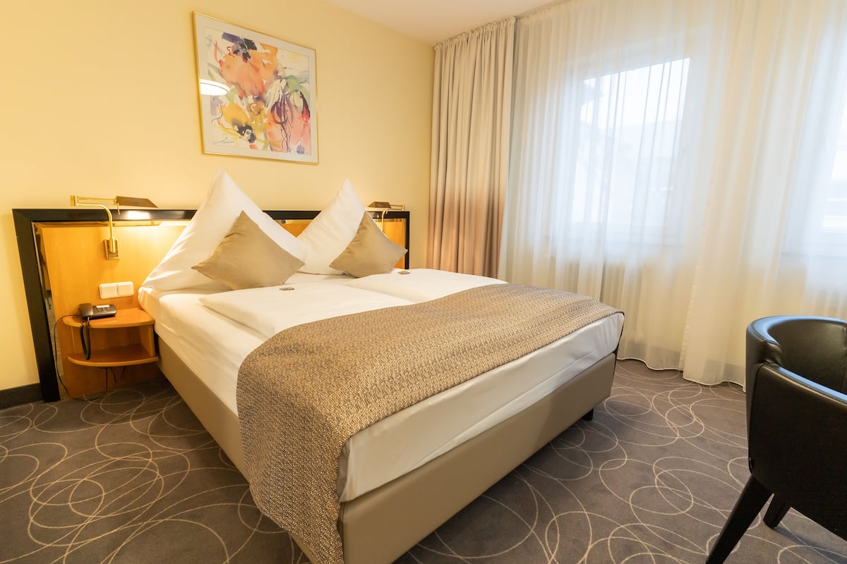 Quarto de hotel com uma cama grande, uma mesa de cabeceira com abajur, uma cadeira e um quadro na parede.