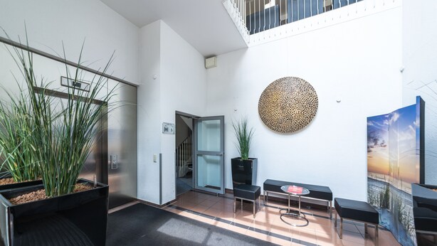 A modern lobby with a glass elevator, a round decorative wall piece, and a seating area with a small table and benches. - Hotel am Stadtgarten (Radolfzell am Bodensee)