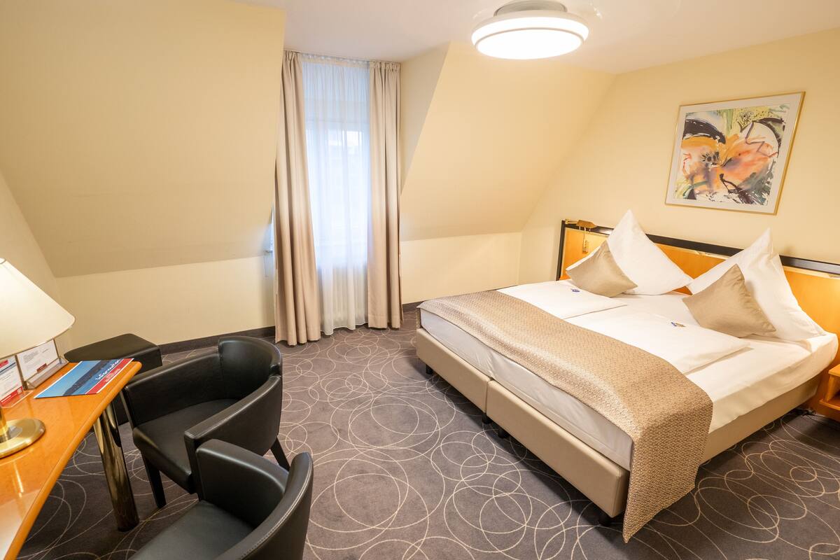 Quarto de hotel com cama, escrivaninha com cadeira, luminária e um quadro na parede.