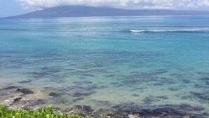 Vista costera con agua turquesa cristalina, costa rocosa y una montaña distante bajo un cielo parcialmente nublado.