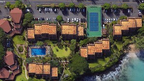 Vista aérea de un complejo turístico costero con varias construcciones tipo bungalow, una piscina y una cancha de tenis.