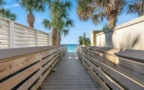 Une promenade en bois menant à une plage bordée de palmiers, sous un ciel bleu et dégagé.