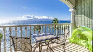A balcony with a table and chairs, overlooking a clear blue sea with boats and a distant mountain. - 6 th floor ocean front condo Outstanding Views! Weekly discounts available  (Lahaina)