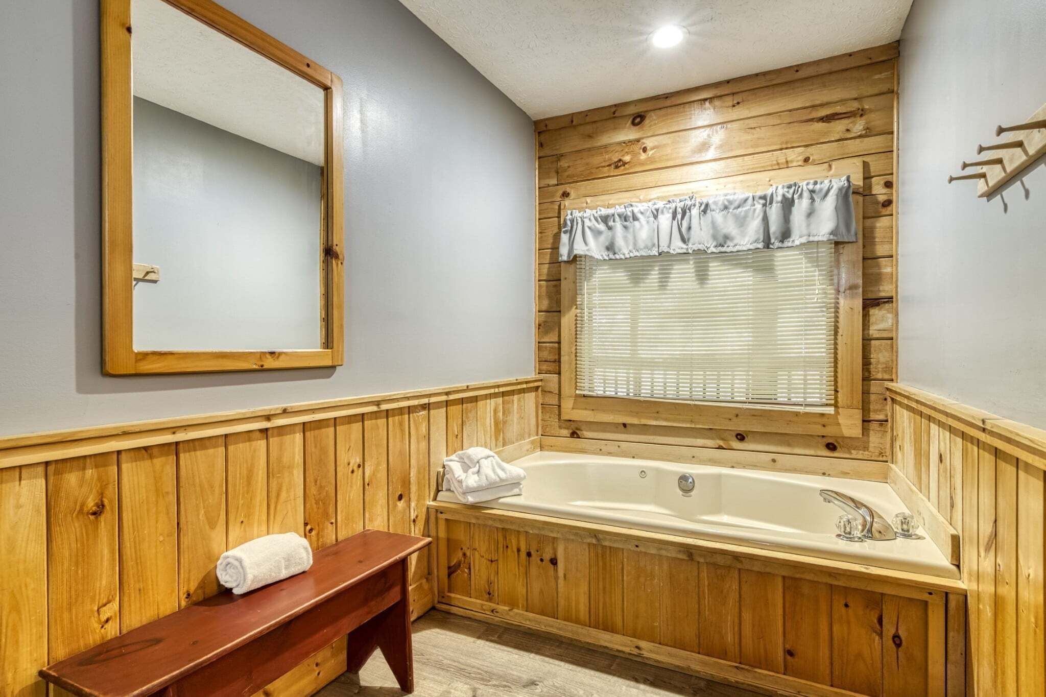 A wooden-paneled bathroom with a large bathtub, a wooden bench, and a window with blinds.