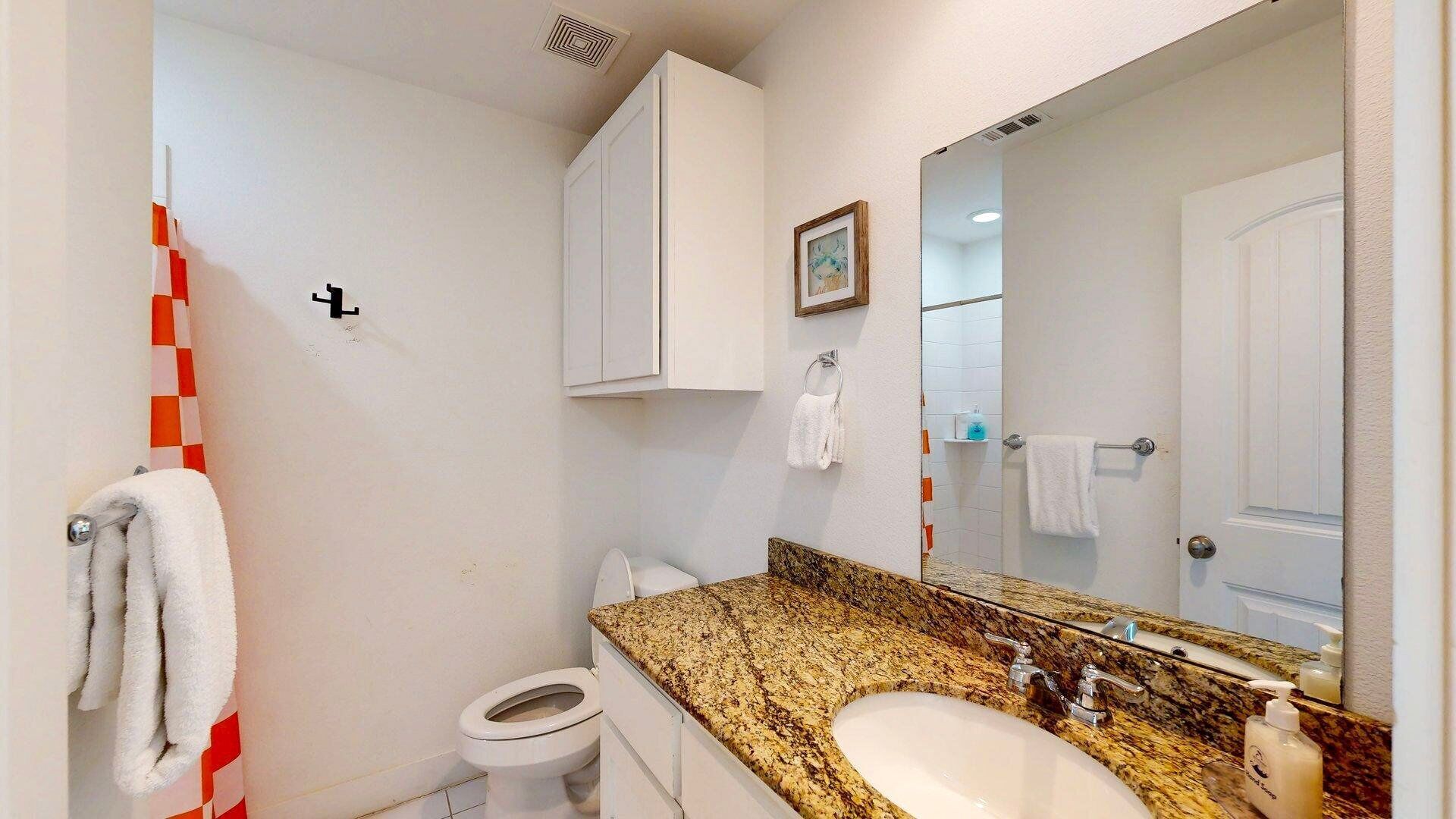 Ein Badezimmer mit Toilette, Waschbecken und Spiegel. Handtücher hängen an der Wand und im Hintergrund ist eine Duschkabine zu sehen.