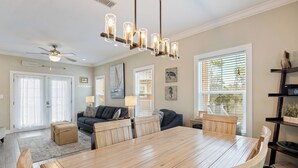 Amplia sala de estar con mesa de comedor de madera, asientos cómodos, ventilador de techo y ventanas grandes con persianas.