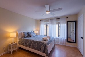 Ein Schlafzimmer mit einem Bett, einer Stehlampe, einem Deckenventilator und einem großen Fenster mit Vorhängen.