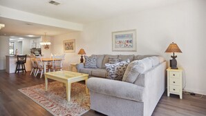 Ein Wohnzimmer mit grauem Sofa, einem Couchtisch und einem Essbereich mit Stühlen und Tisch. An der Wand hängt ein Gemälde und auf einem Beistelltisch steht eine Lampe.