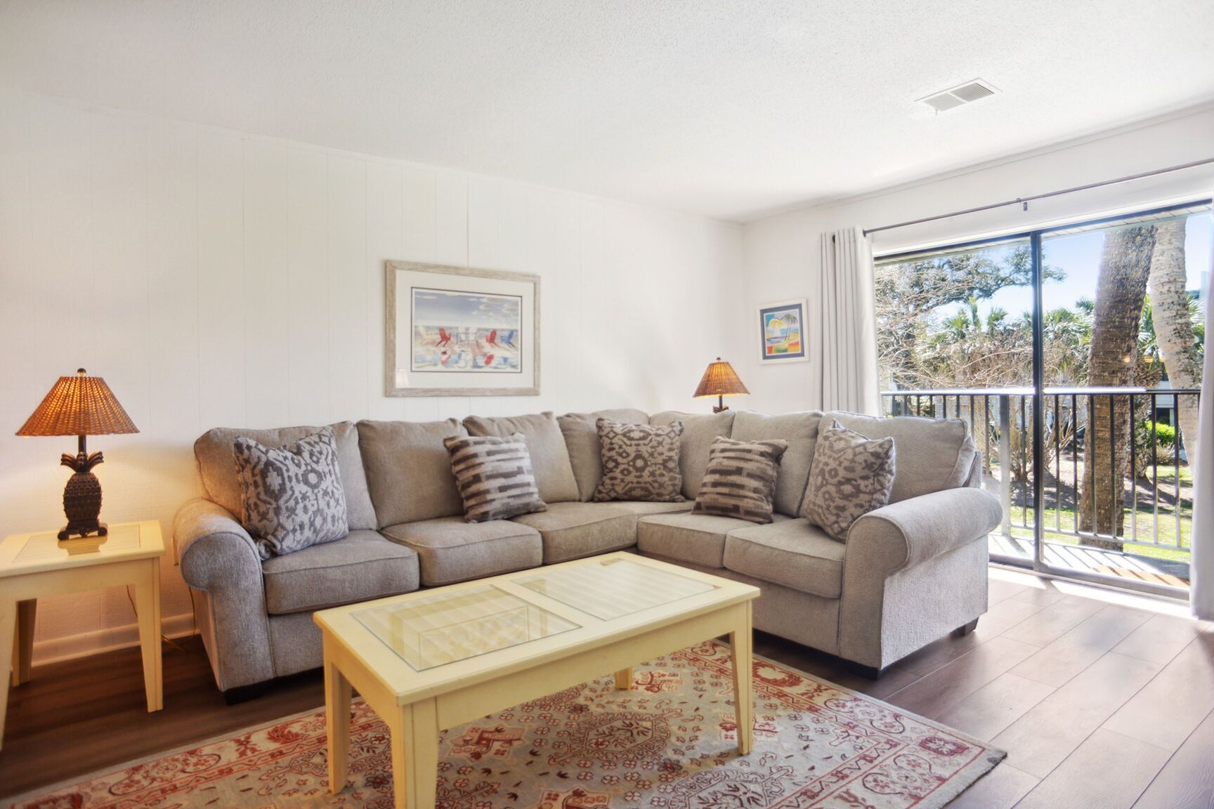 A living room with a sofa, two side tables, a coffee table, a patterned rug, and a balcony with a view of trees.