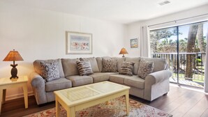 Ein Wohnzimmer mit einer Couch, zwei Beistelltischen, einem Couchtisch, einem gemusterten Teppich und einem Balkon mit Blick auf Bäume.