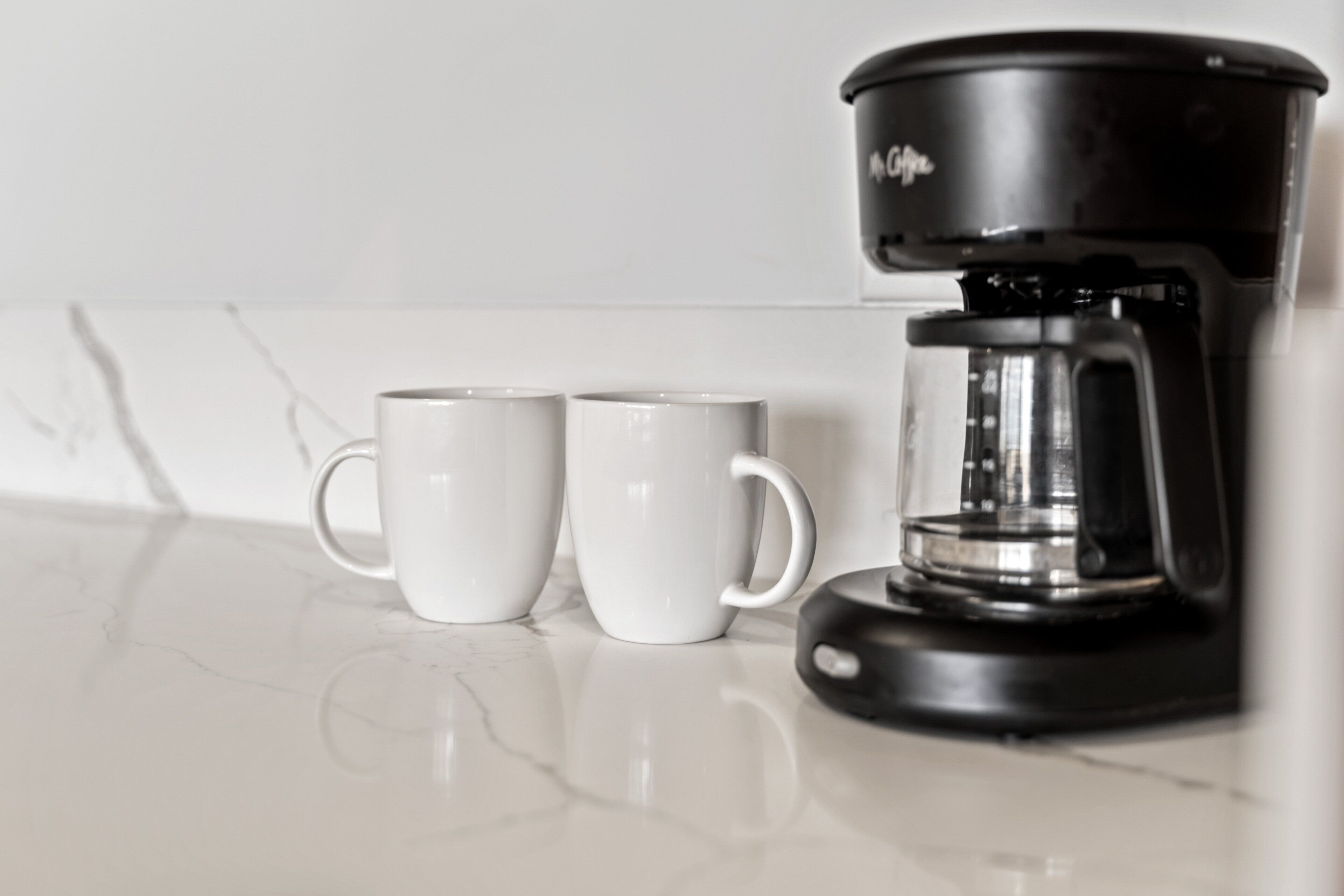 Una cafetera con dos tazas blancas sobre una encimera de mármol.