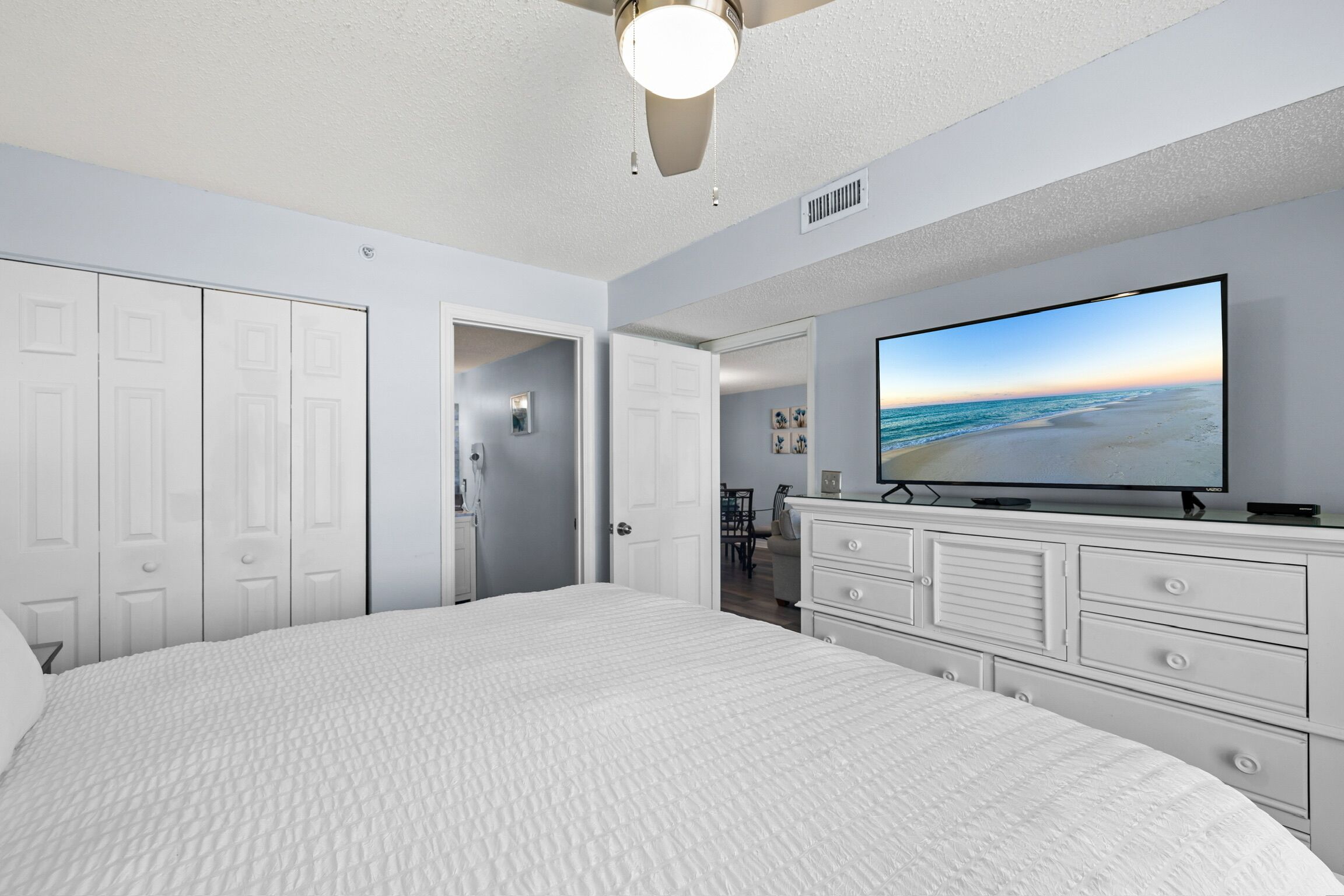 A bedroom with a large bed, a ceiling fan, a TV on a dresser, and a view of the beach.
