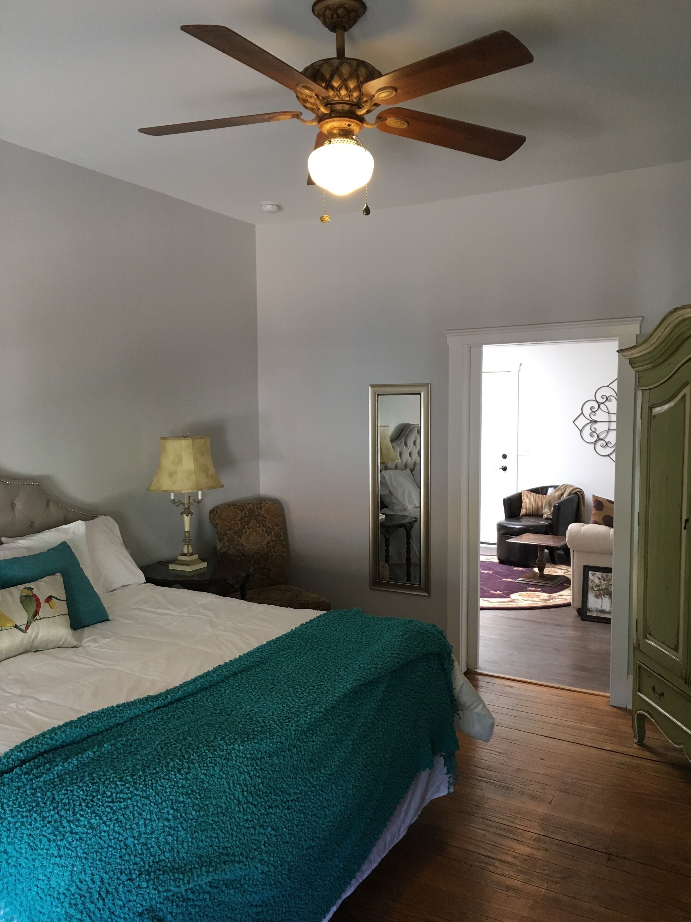 A bedroom with a bed, a ceiling fan, a nightstand with a lamp, a chair, and a mirror.