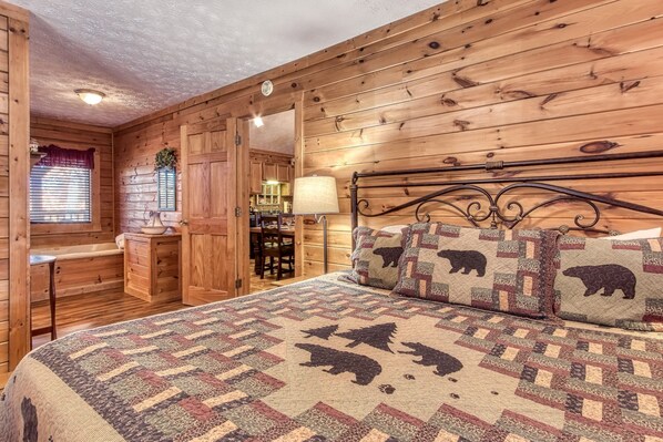 A cozy cabin-style room with wooden walls, a bed with a quilted blanket, a wooden bench, a small table, and a window with blinds.