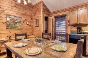 Cozy cabin interior with a dining table for four, wooden walls, and a kitchen area featuring a coffee maker and bread basket.