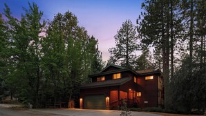 A two-story log cabin with a garage, surrounded by tall trees. - Luxury! Pool Table! Hot Tub! Pet Friendly! Covered Deck! 4 Master Suites! Log Features! (Big Bear City)