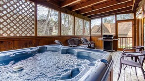 A covered outdoor hot tub area with wooden decking, wicker chairs, and a barbecue grill.