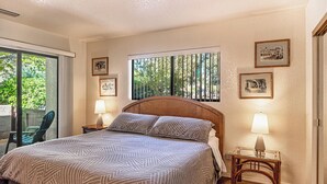 A bedroom with a large bed, two bedside tables with lamps, a window with vertical blinds, and framed pictures on the wall.