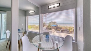 Un comedor moderno con una mesa de vidrio, sillas blancas y vista a la playa a través de ventanas amplias.